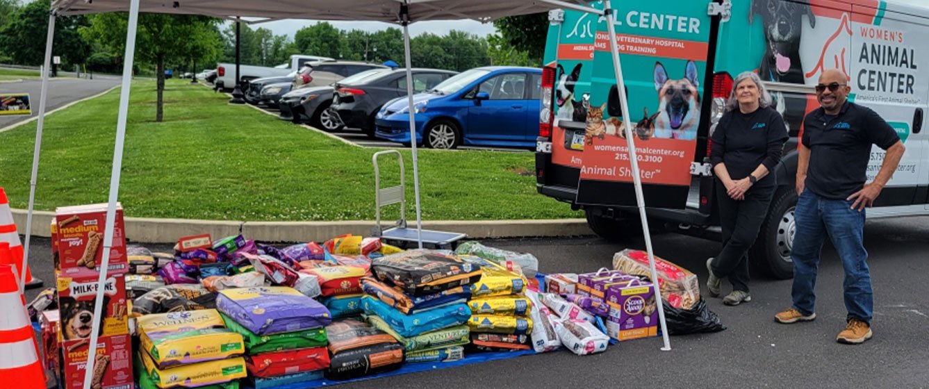 Two people in black shirts stand to the right of a stack of dog and cat food bags in a parking lot.