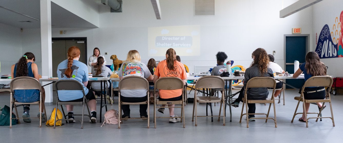 Teens sit at a table in a large room while a presentation occurs in front of them