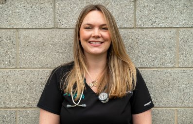 A white women in black scrubs with a stethoscope around her neck stands in front of a gray brick wall. She is smiling.