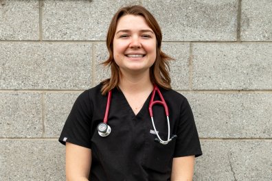 A white woman in black scrubs with a stethoscope around her neck stands in front of a gray brick wall. She is smiling.