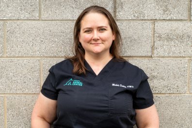 A white women in black scrubs stands in front of a gray brick wall. She is smiling.