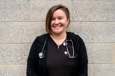 A woman in black scrubs with a stethoscope around her neck smiles in front of a gray brick wall.